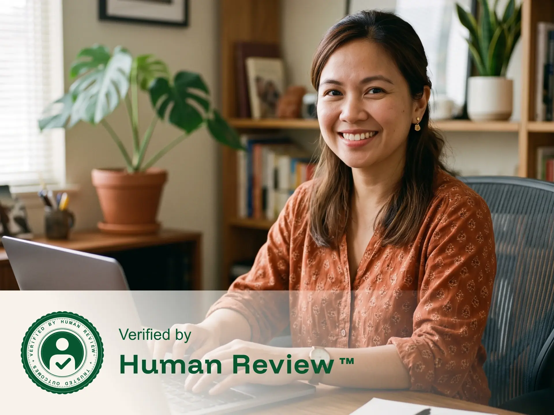Smiling woman working on laptop at a desk with houseplants and bookshelves behind her.