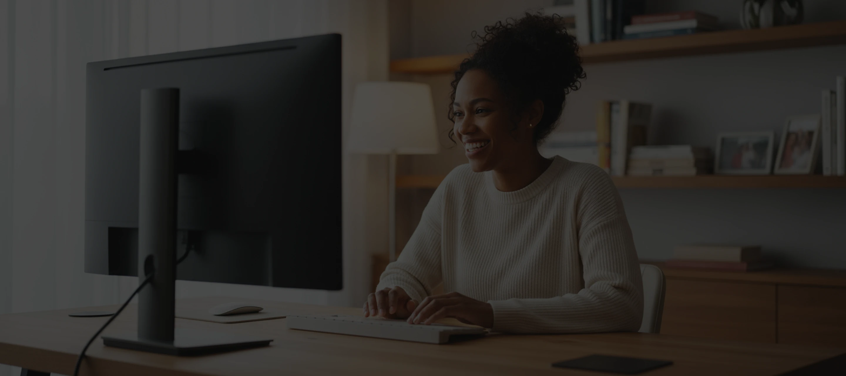 Smiling woman typing on a keyboard at a desk with a monitor and shelves in the background.