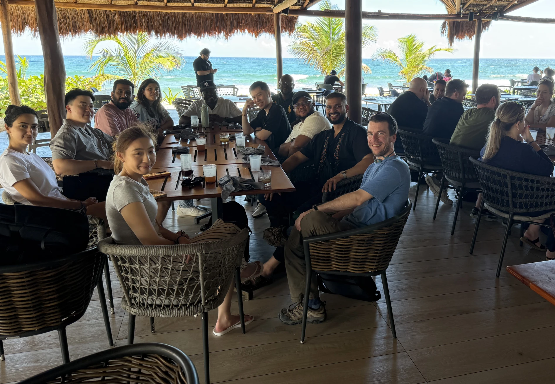 Group of people seated around a table at a seaside restaurant with ocean and palm trees in the background.