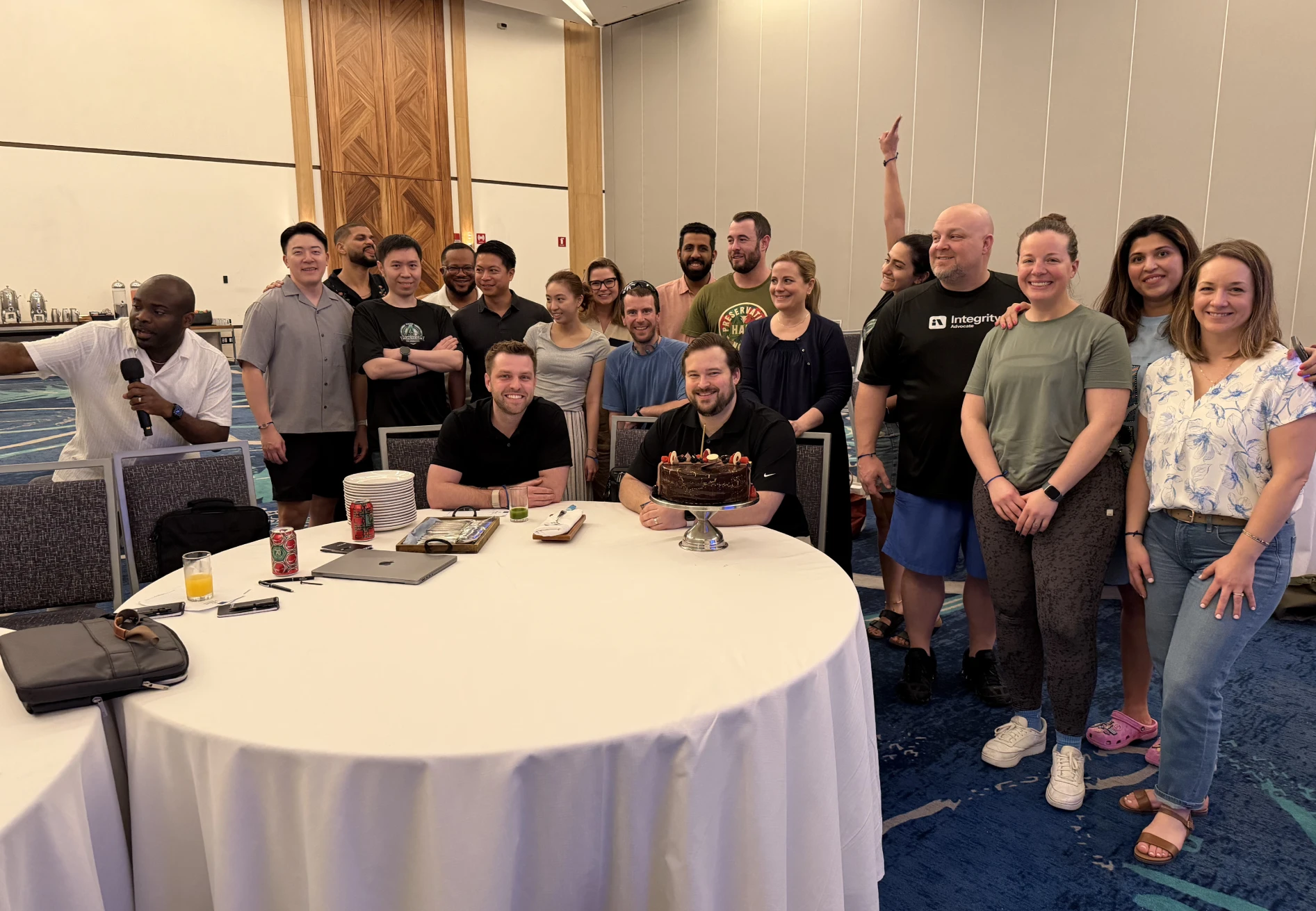 Group of people smiling around table with chocolate cake in a conference room setting.