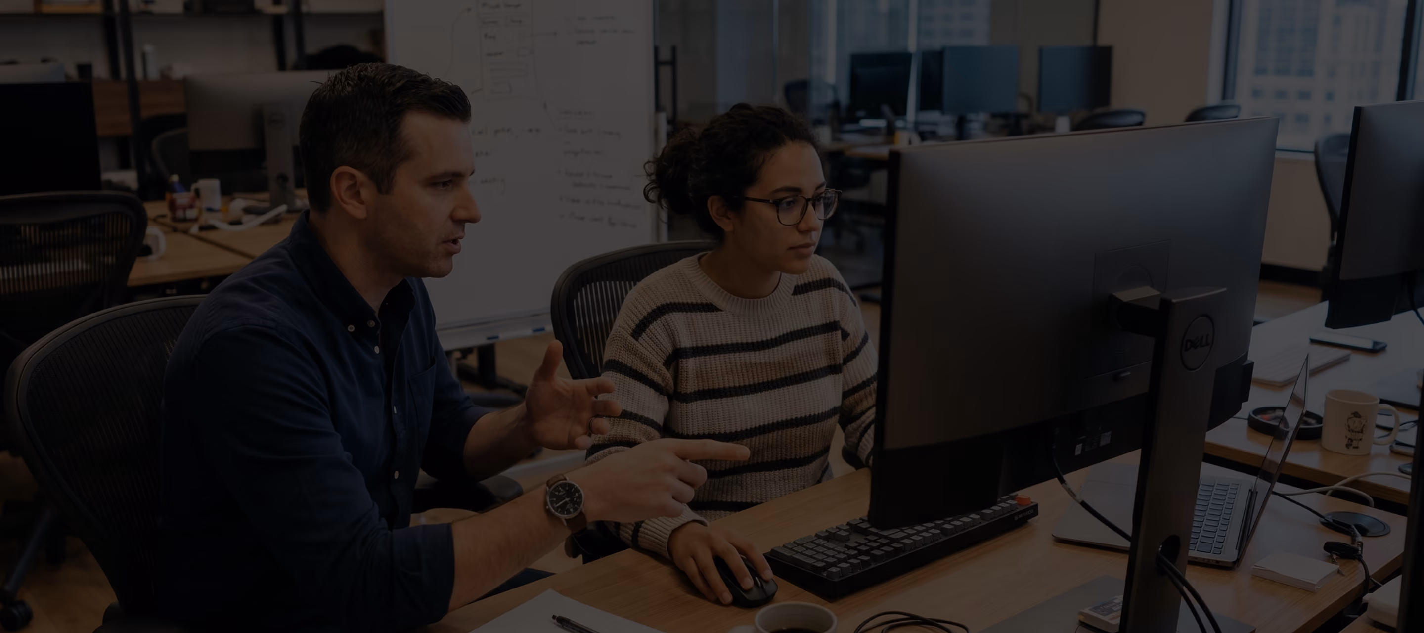Two coworkers discuss work while looking at a computer screen in an office with desks and monitors.