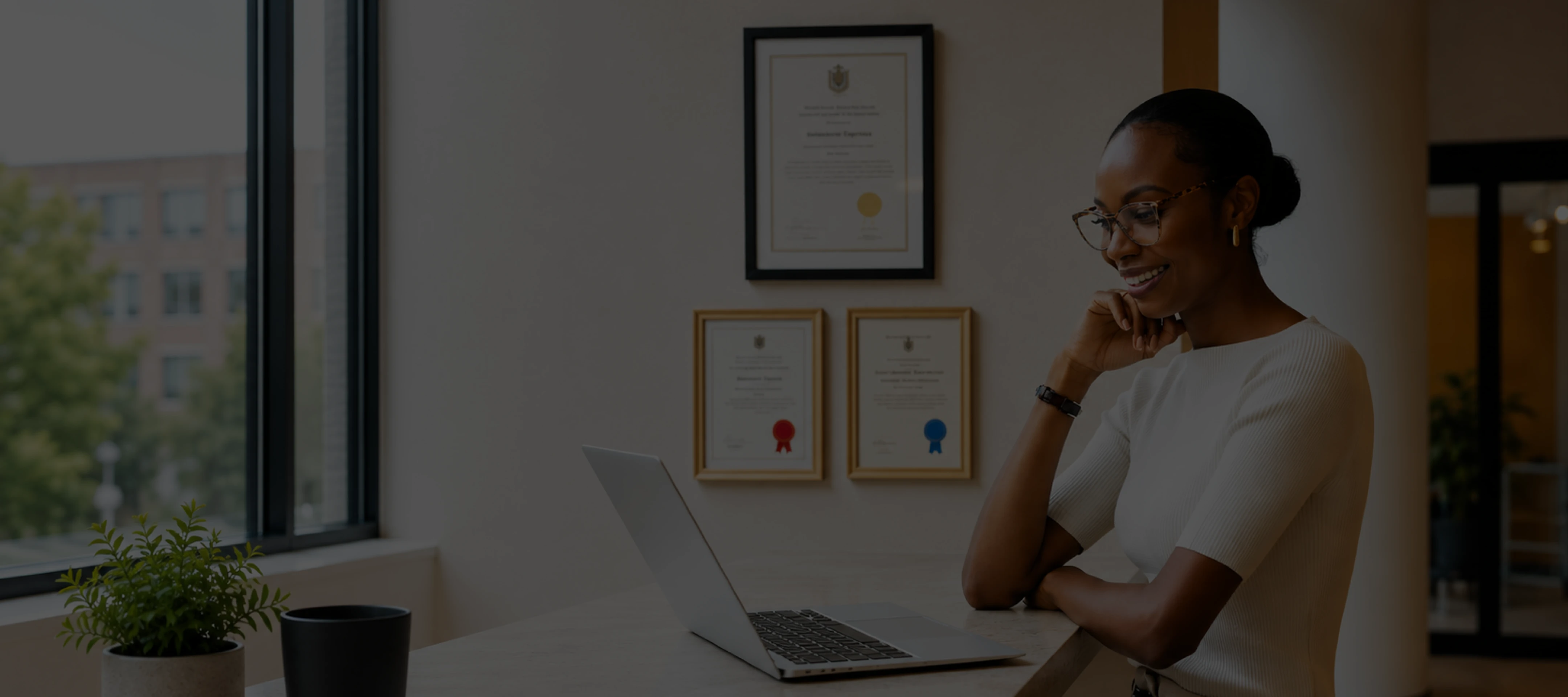 Woman wearing glasses smiling while working on a laptop at a counter near certificates on the wall.
