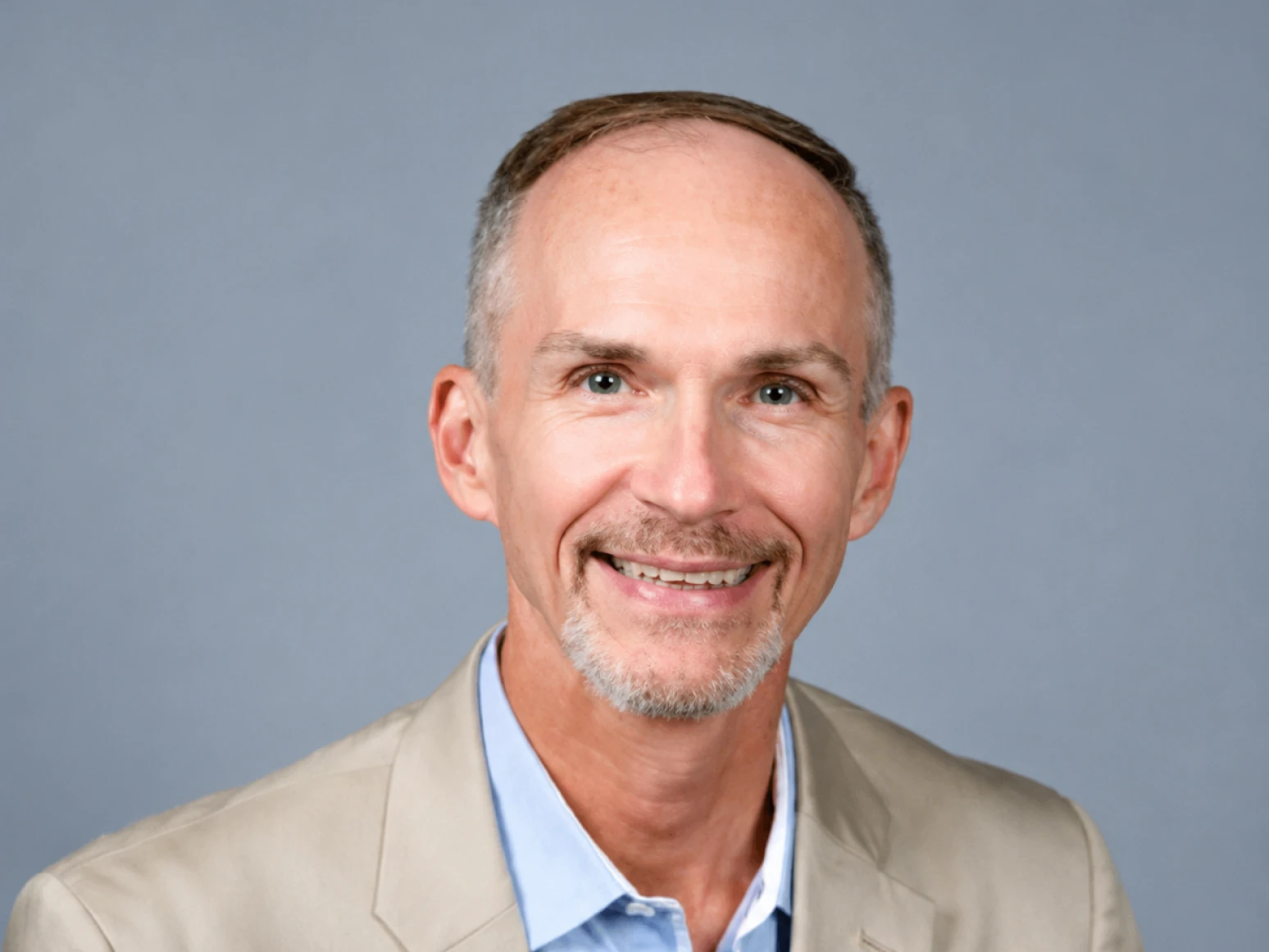 Smiling middle-aged man with short gray hair and beard wearing a beige blazer and blue shirt.