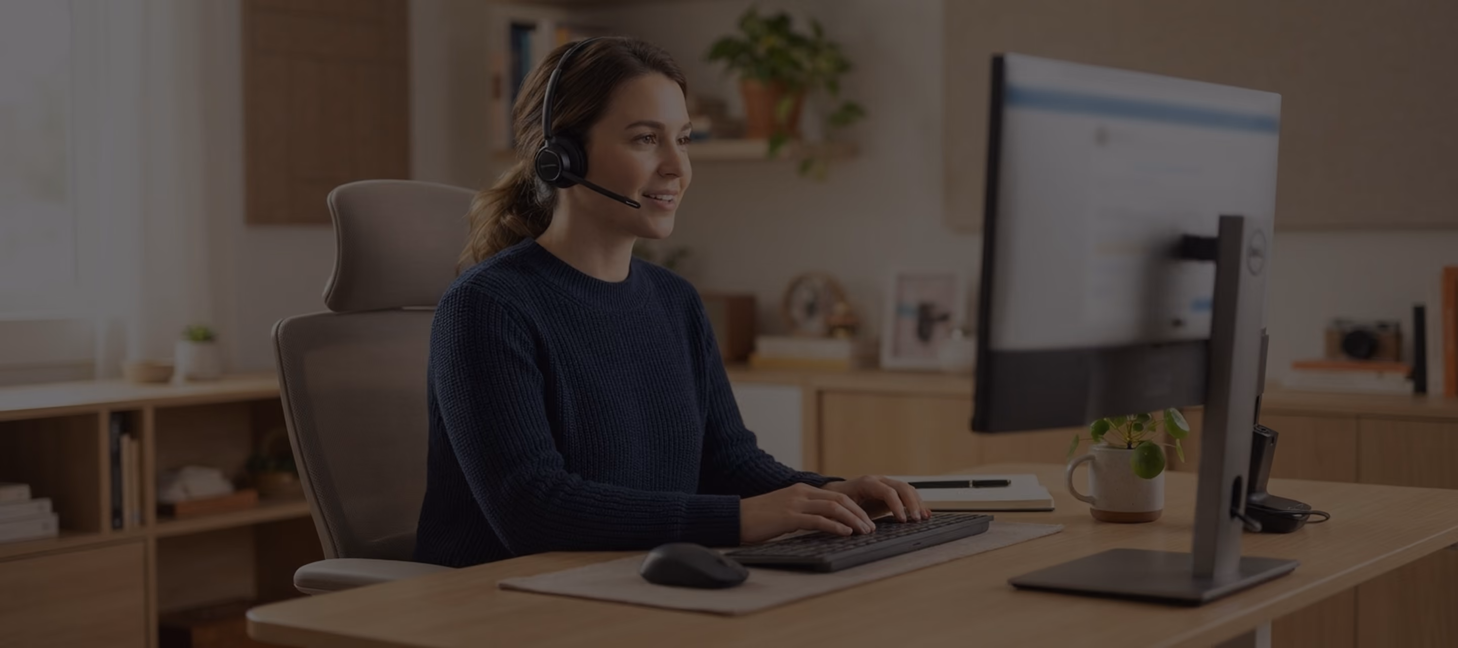 Woman wearing headset typing on keyboard while looking at desktop monitor in home office.
