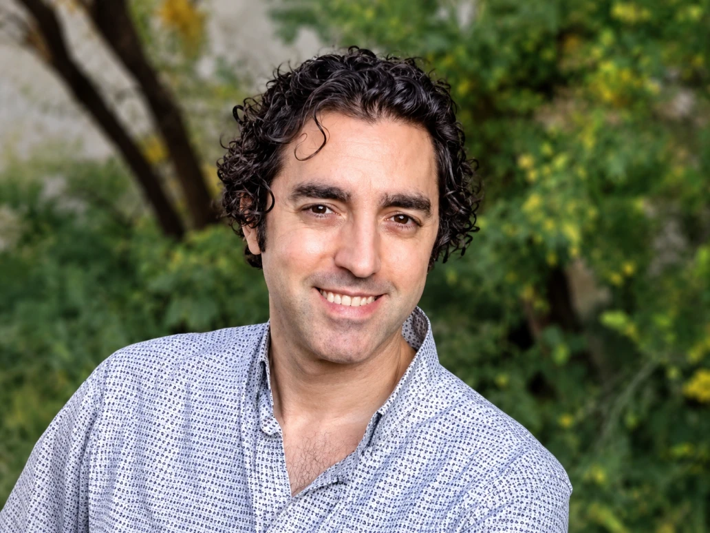 Smiling man wearing a patterned shirt standing outdoors with green foliage behind him.