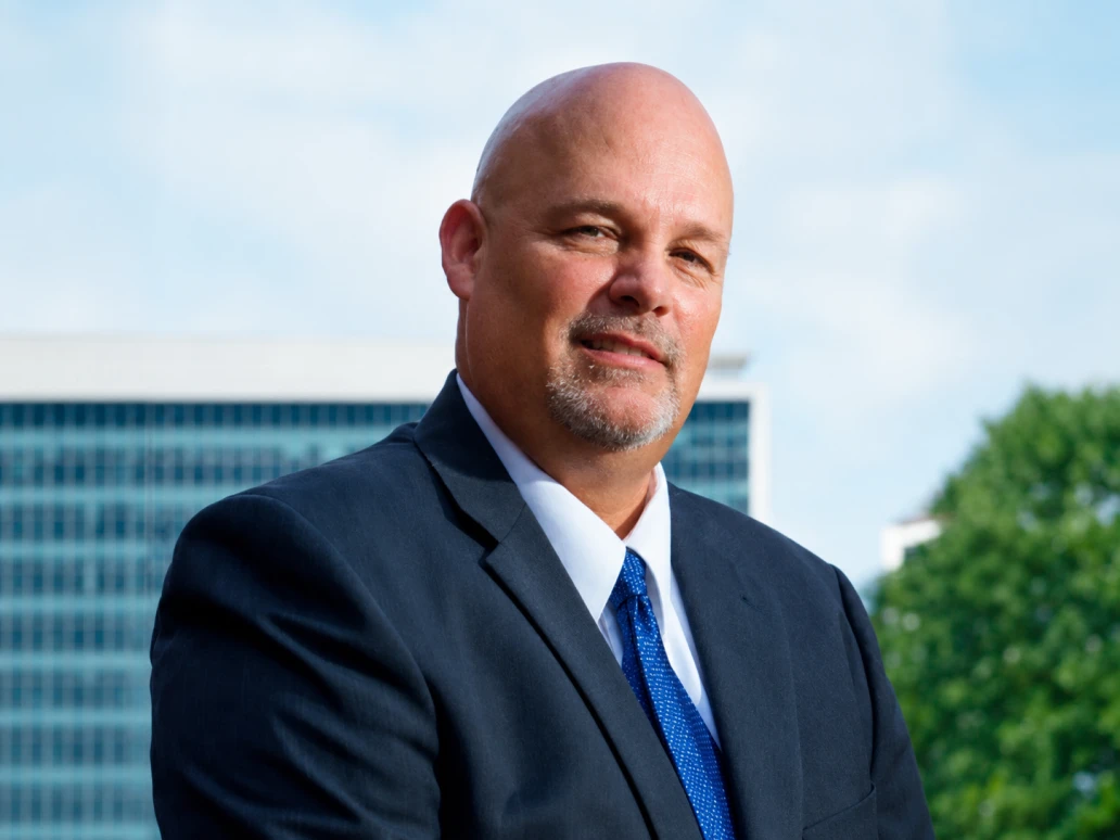 Man in a dark suit and blue tie outdoors with a blurred building and tree in the background.