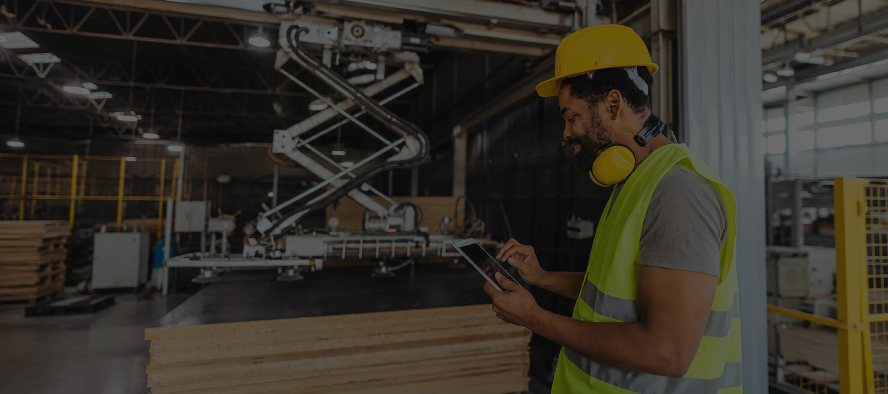 Factory worker in yellow helmet and vest uses tablet near automated machinery and stacked wood panels.