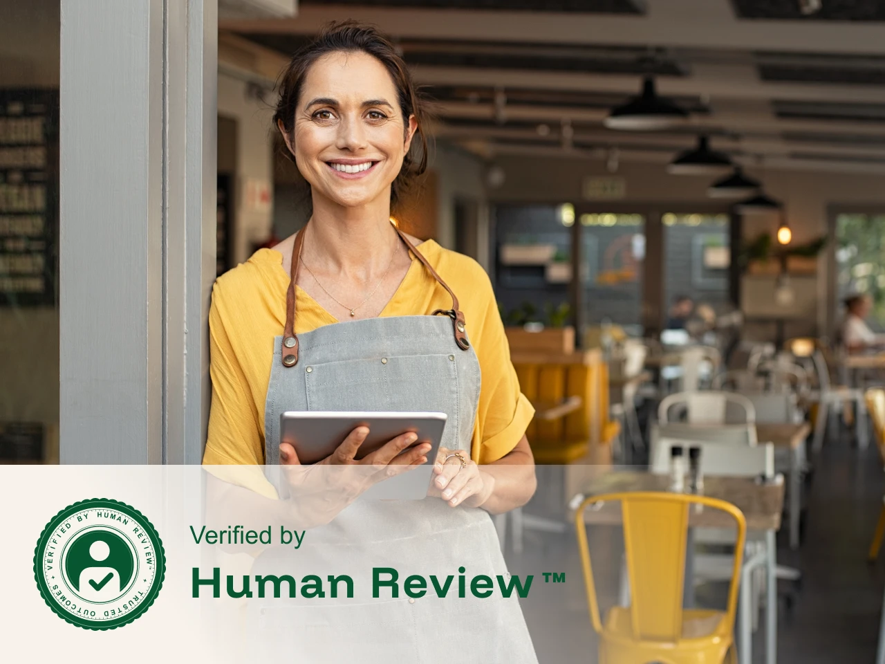 Smiling woman in apron holding tablet inside a bright, modern restaurant dining area.