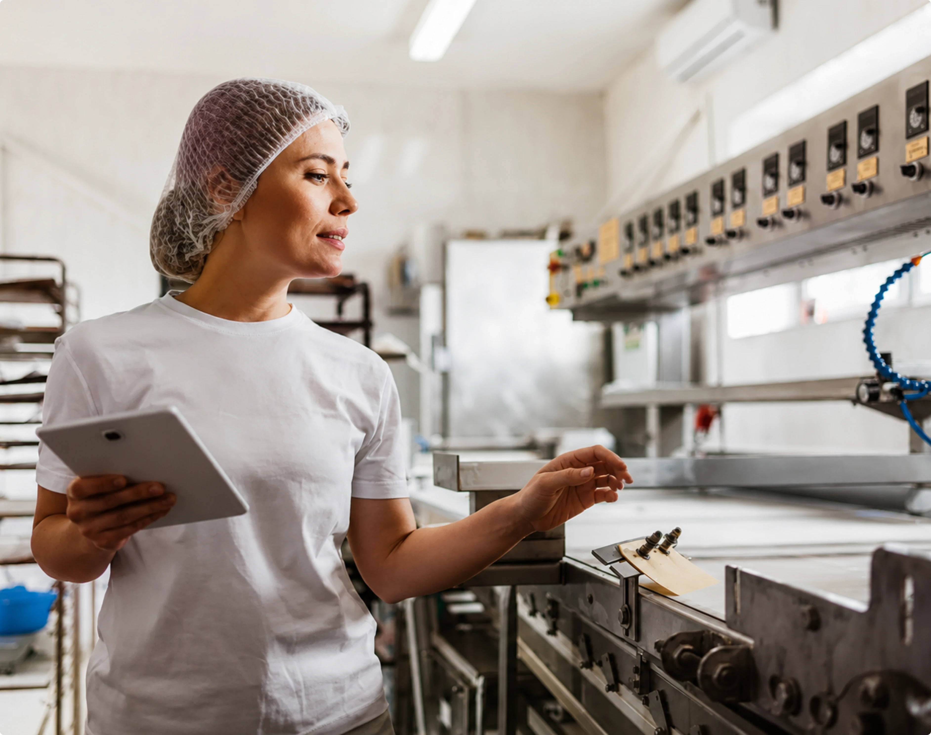 Woman in hairnet and white shirt checking industrial food processing equipment holding a tablet.