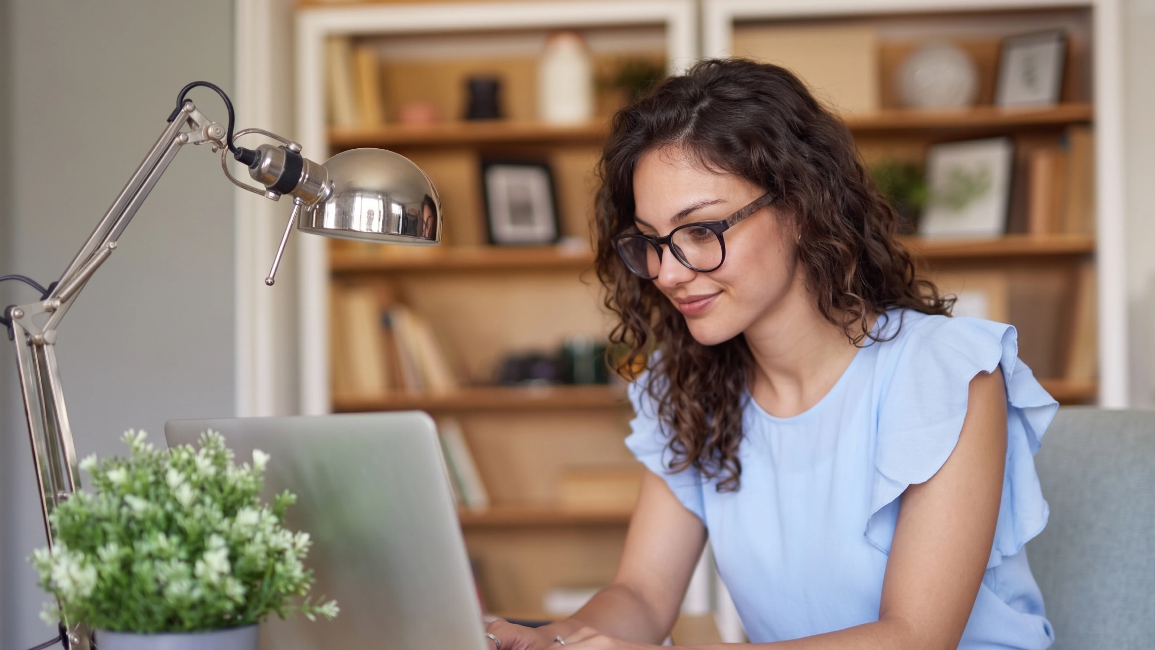 Woman with glasses working on a laptop at a desk with a plant and lamp nearby.