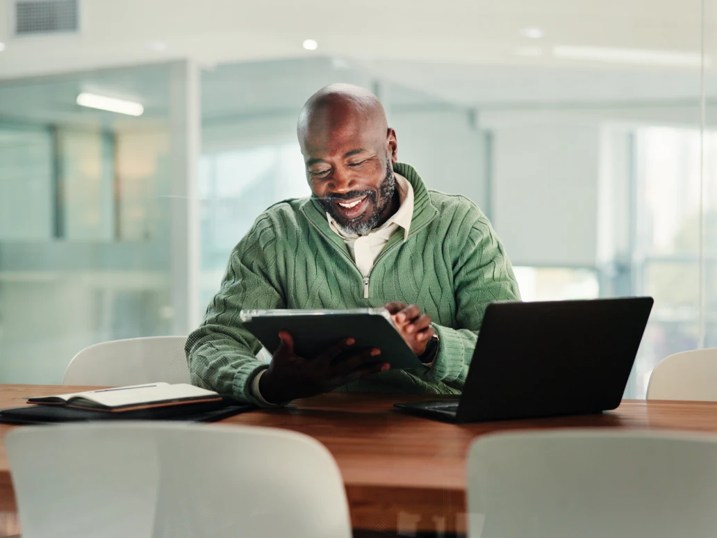 Smiling man in green sweater using a tablet at a table with a laptop and notebook nearby.