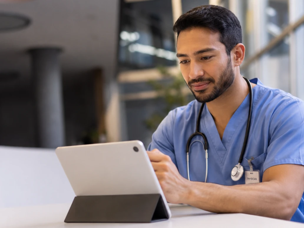 Male doctor in scrubs with stethoscope using tablet at a white desk in a medical facility.