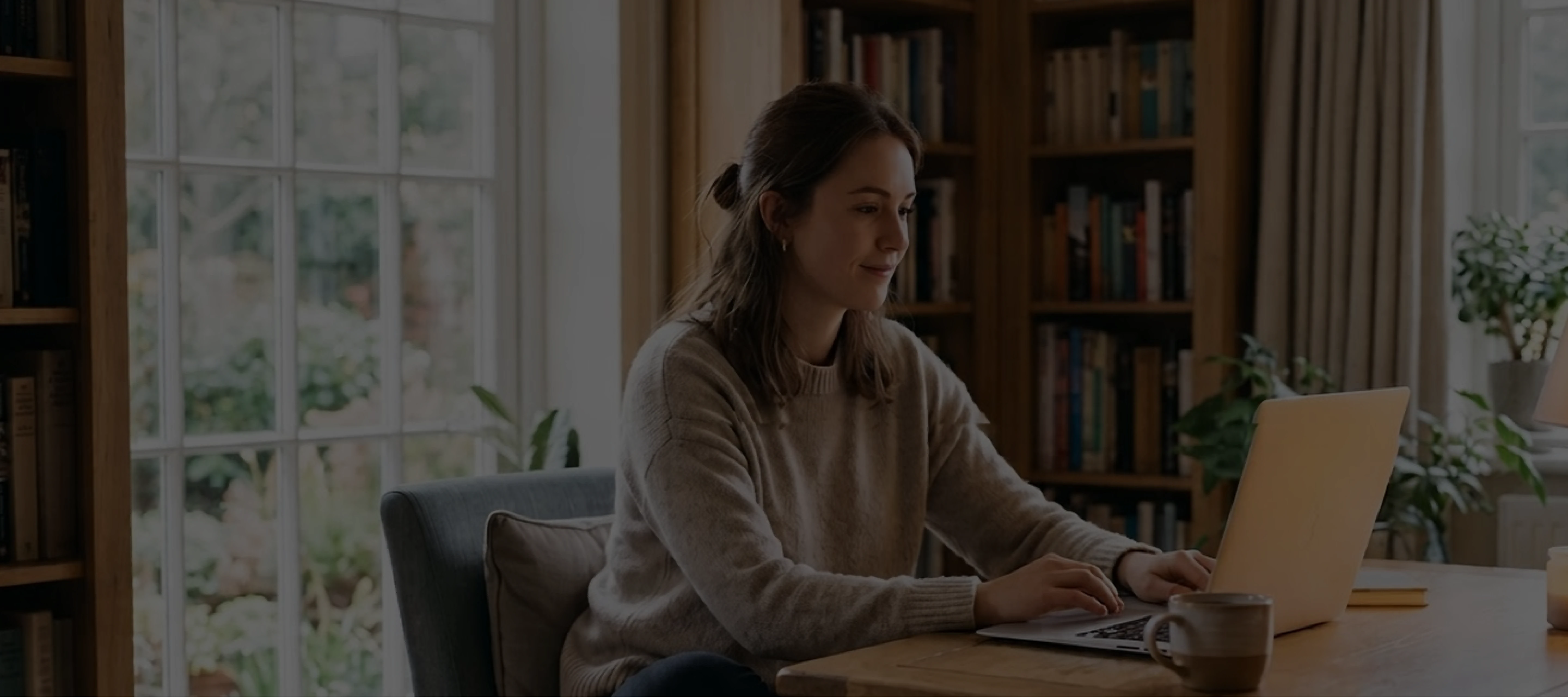 Woman working on laptop at wooden table in cozy room with large window and bookshelves.