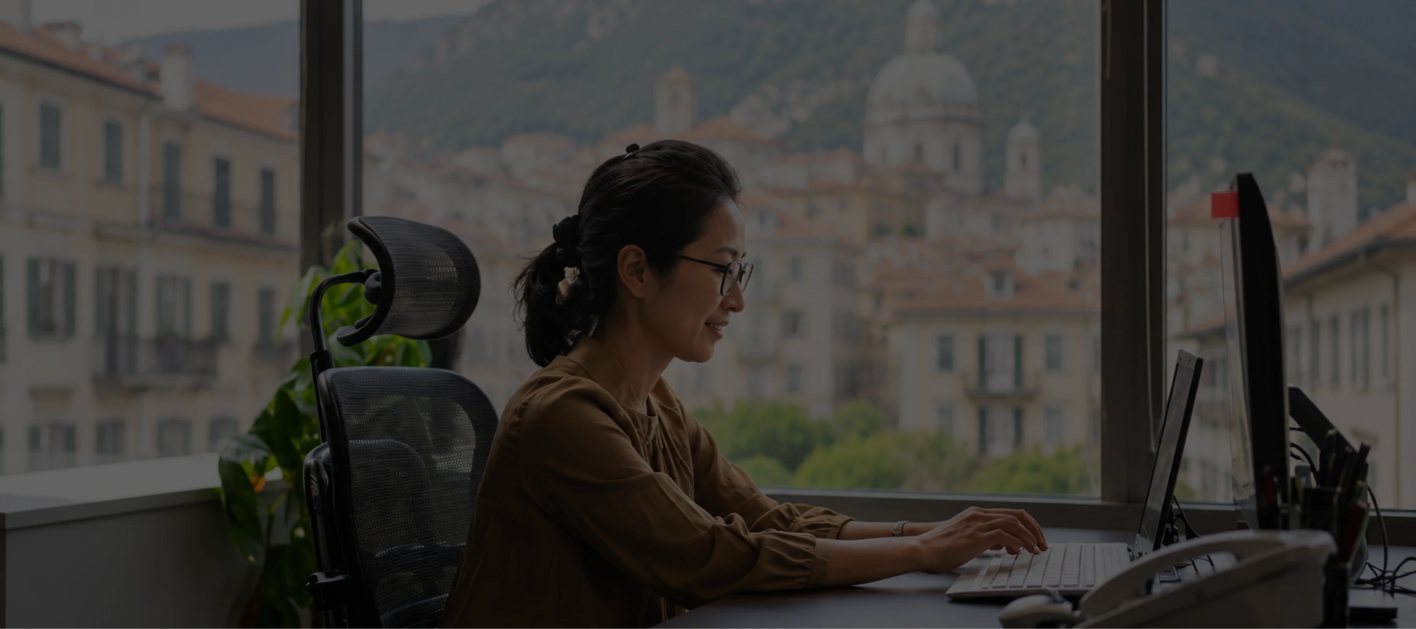 Woman with glasses typing on a laptop at a desk in front of a window showing a cityscape with historic buildings.