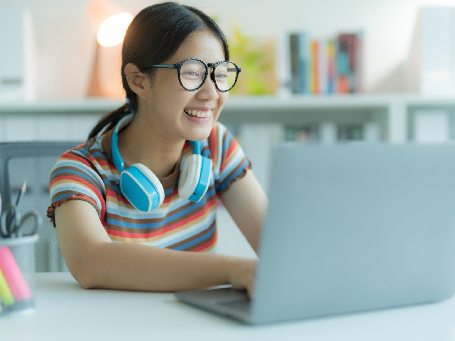 Smiling young woman with glasses and blue headphones using laptop at a desk.