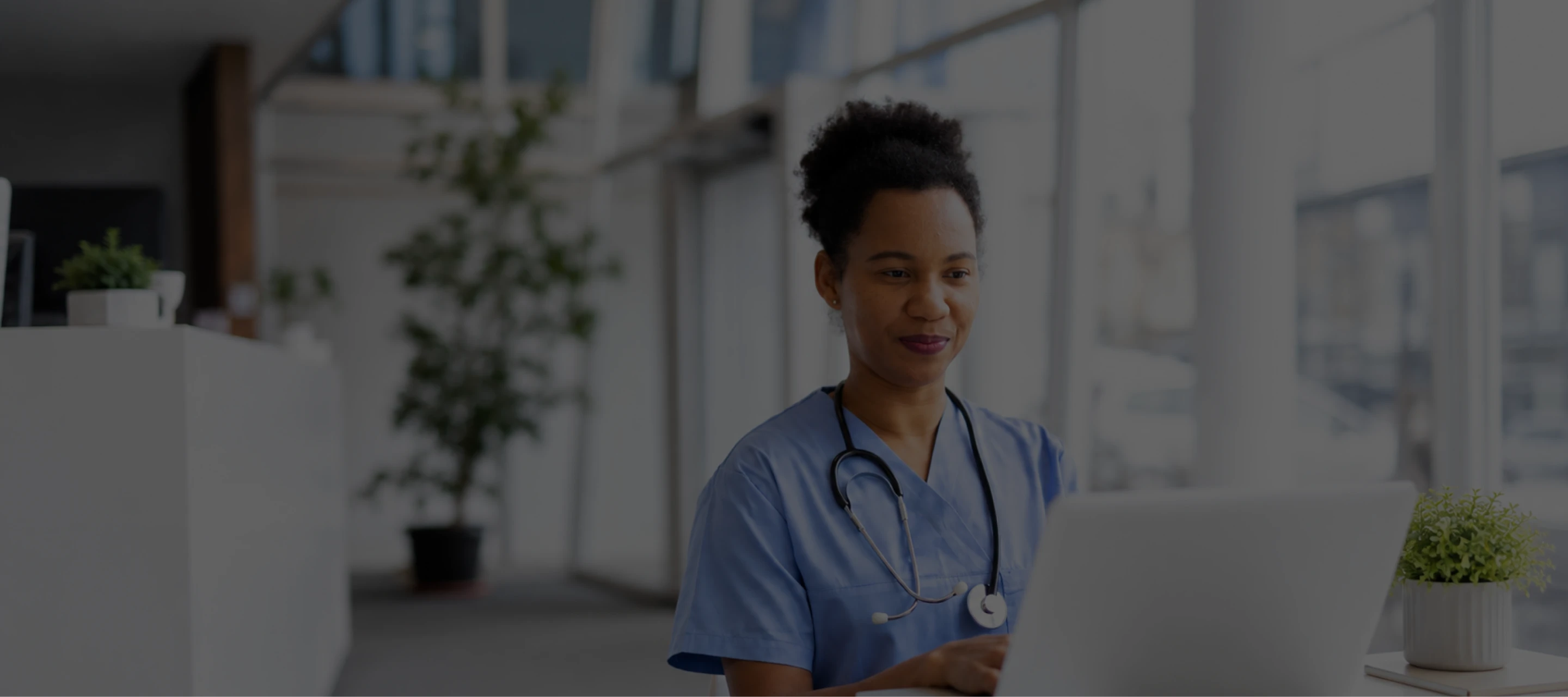 Healthcare professional in blue scrubs using a laptop in a bright modern office.