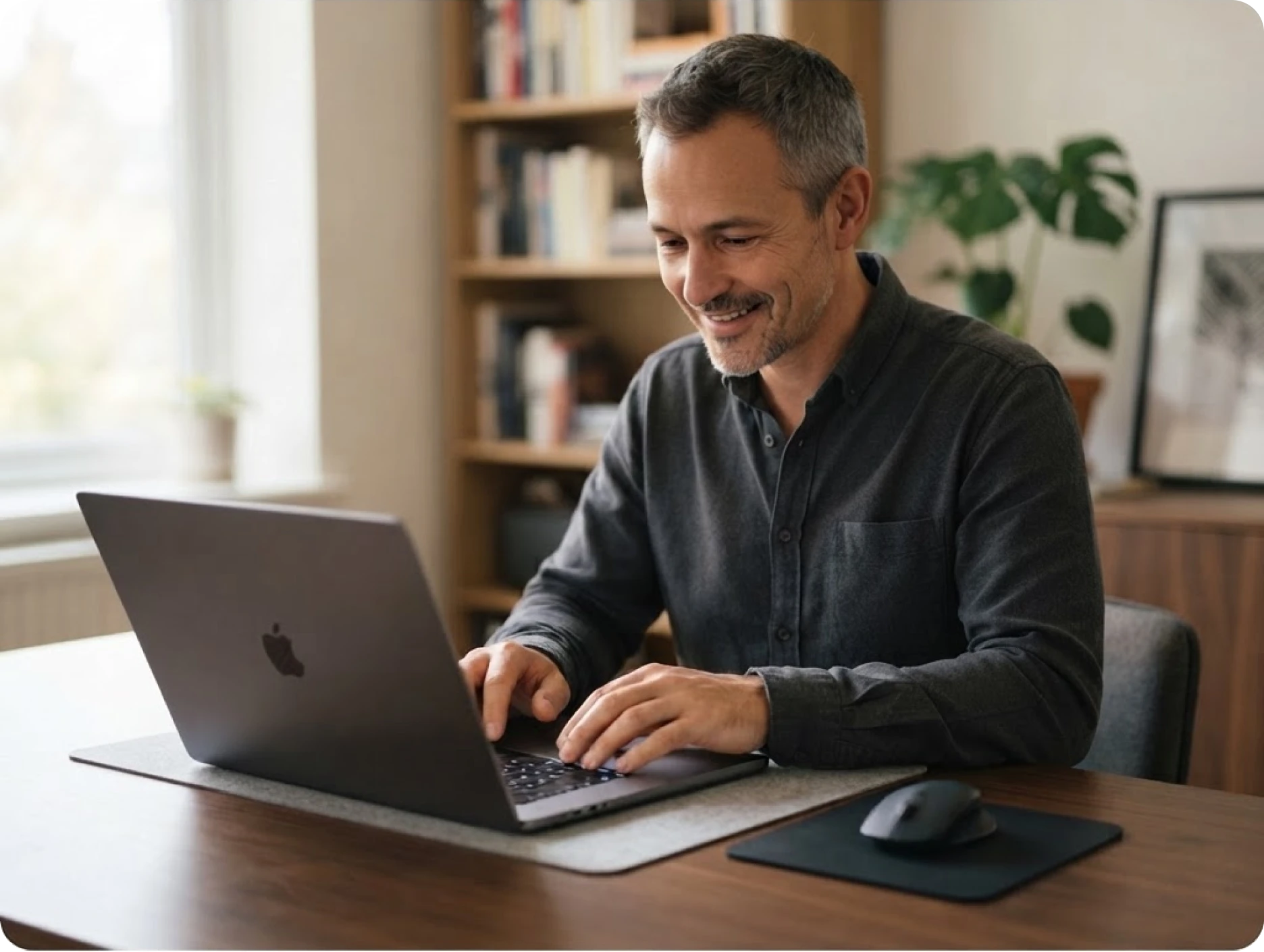 Smiling man typing on a laptop in a cozy room with bookshelves and plants in the background.