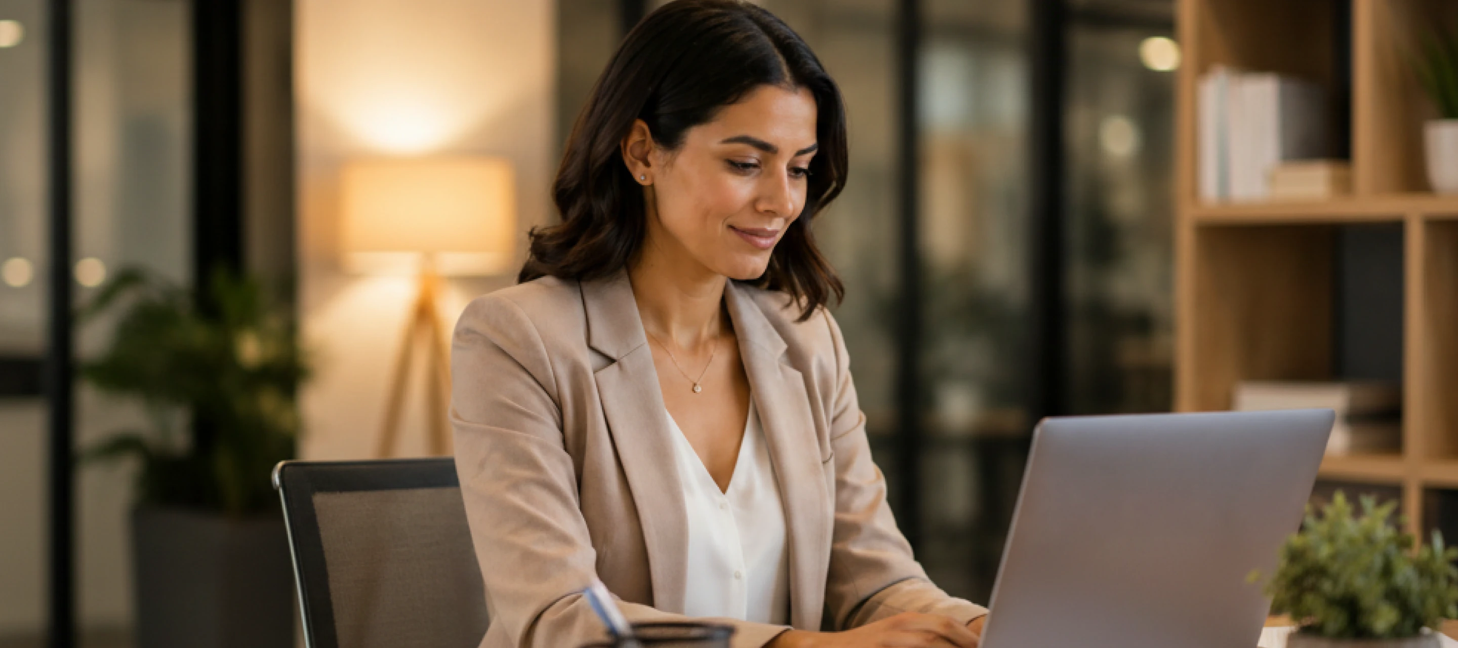 Woman in beige blazer working on a laptop at a desk in a modern office.