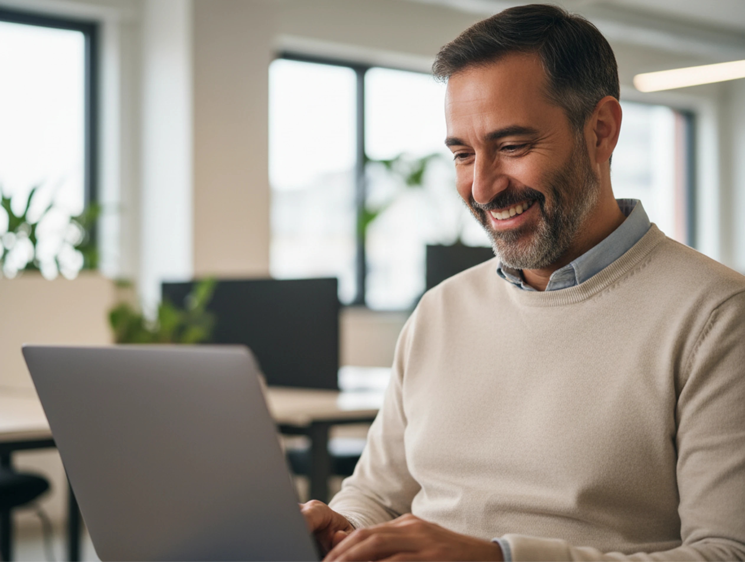Smiling man with beard working on a laptop in a bright modern office with plants in background.