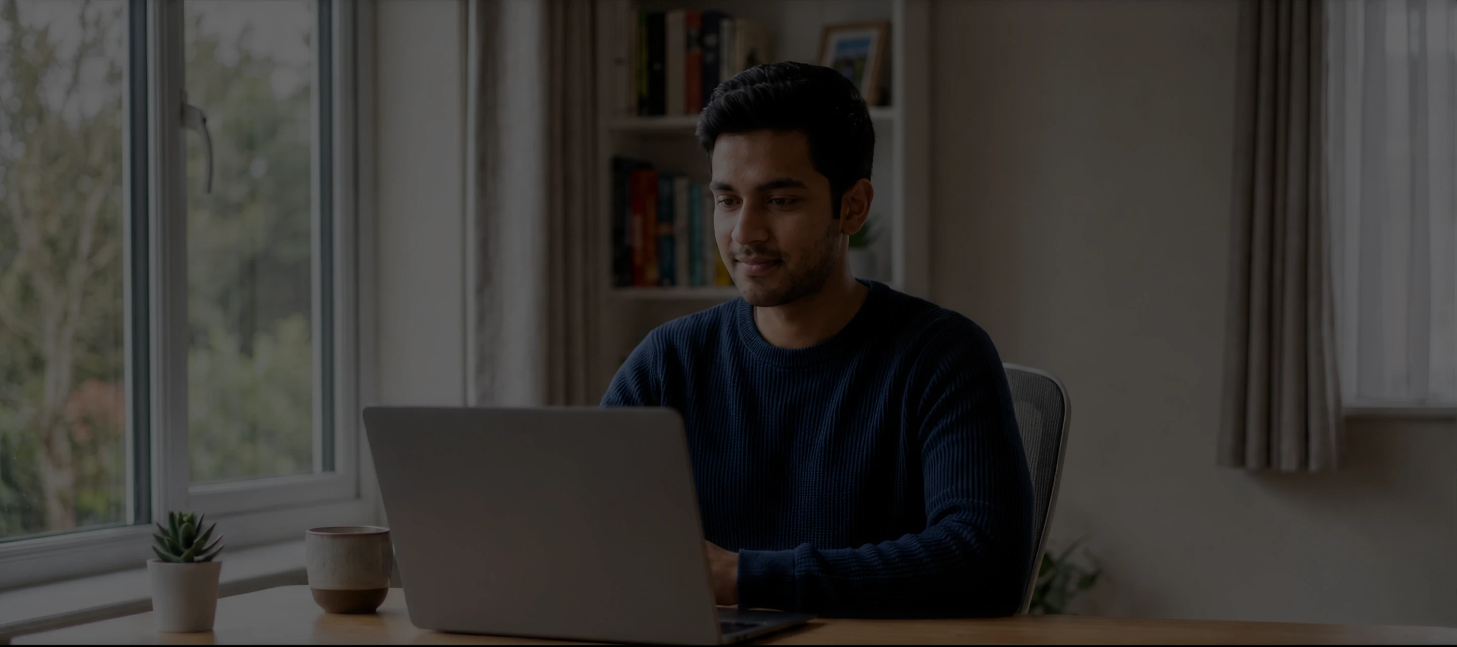 Man in a blue sweater working on a laptop at a desk near a window with plants.