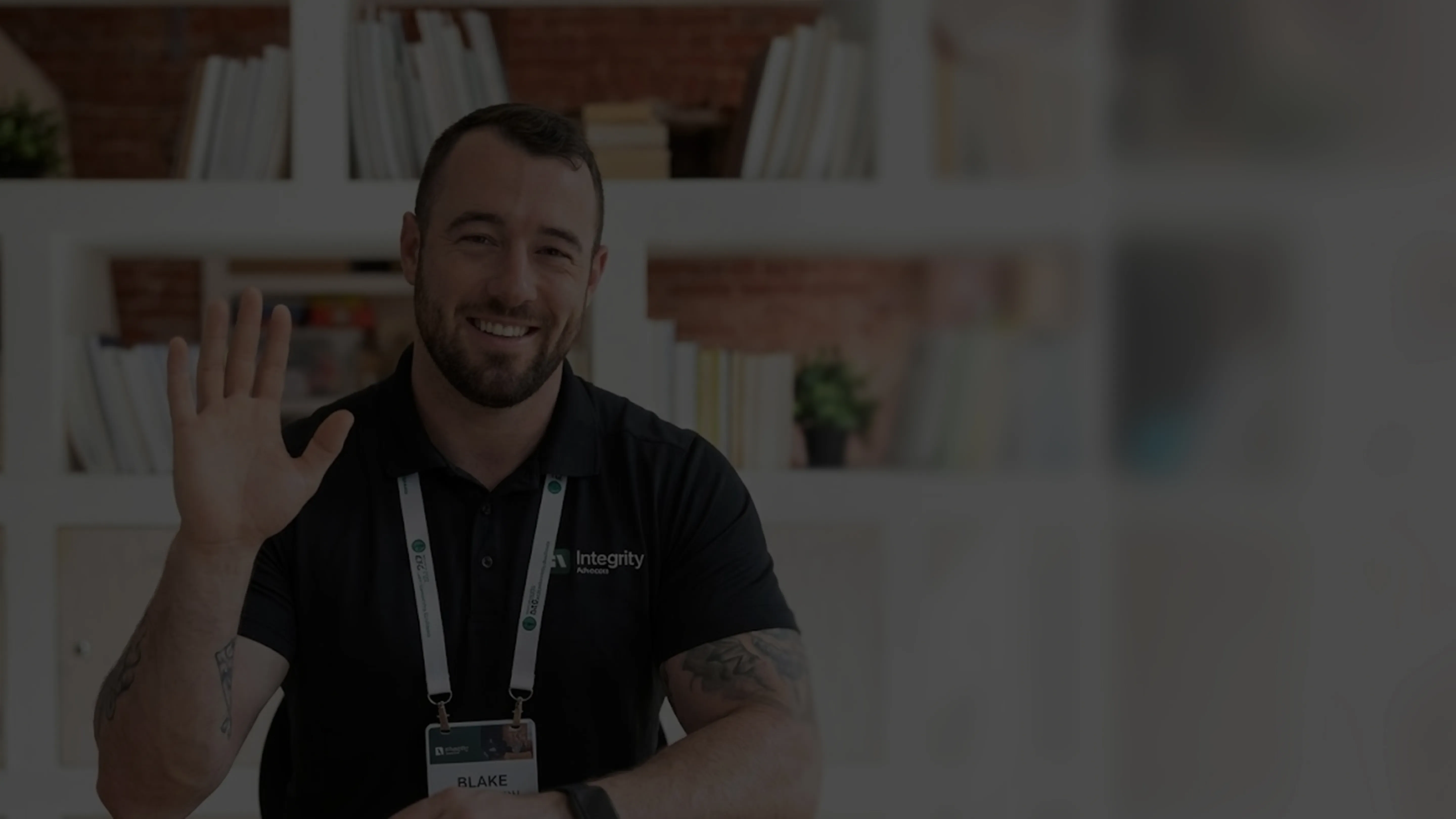 Smiling man with beard waving, wearing a black polo and name badge in a room with shelves behind him.