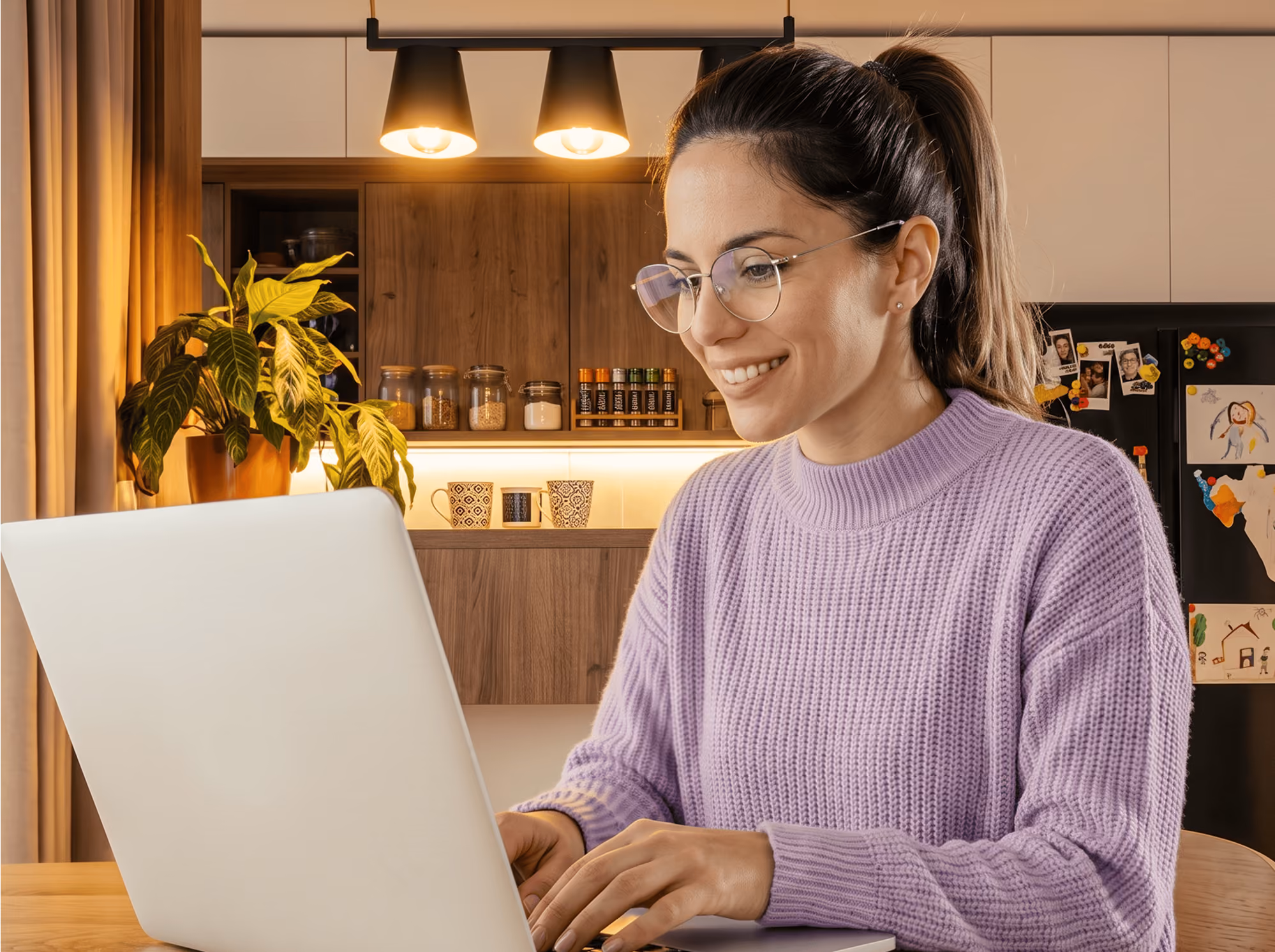 Smiling woman in glasses and purple sweater working on a laptop in a warmly lit kitchen.