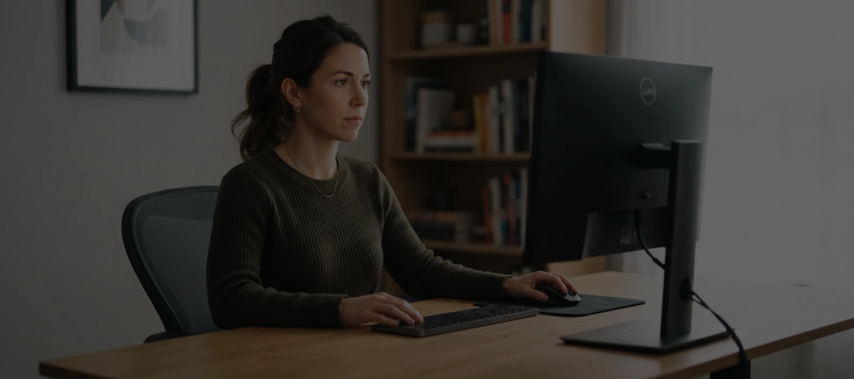 Woman in a dark sweater working on a desktop computer at a wooden desk in an office.