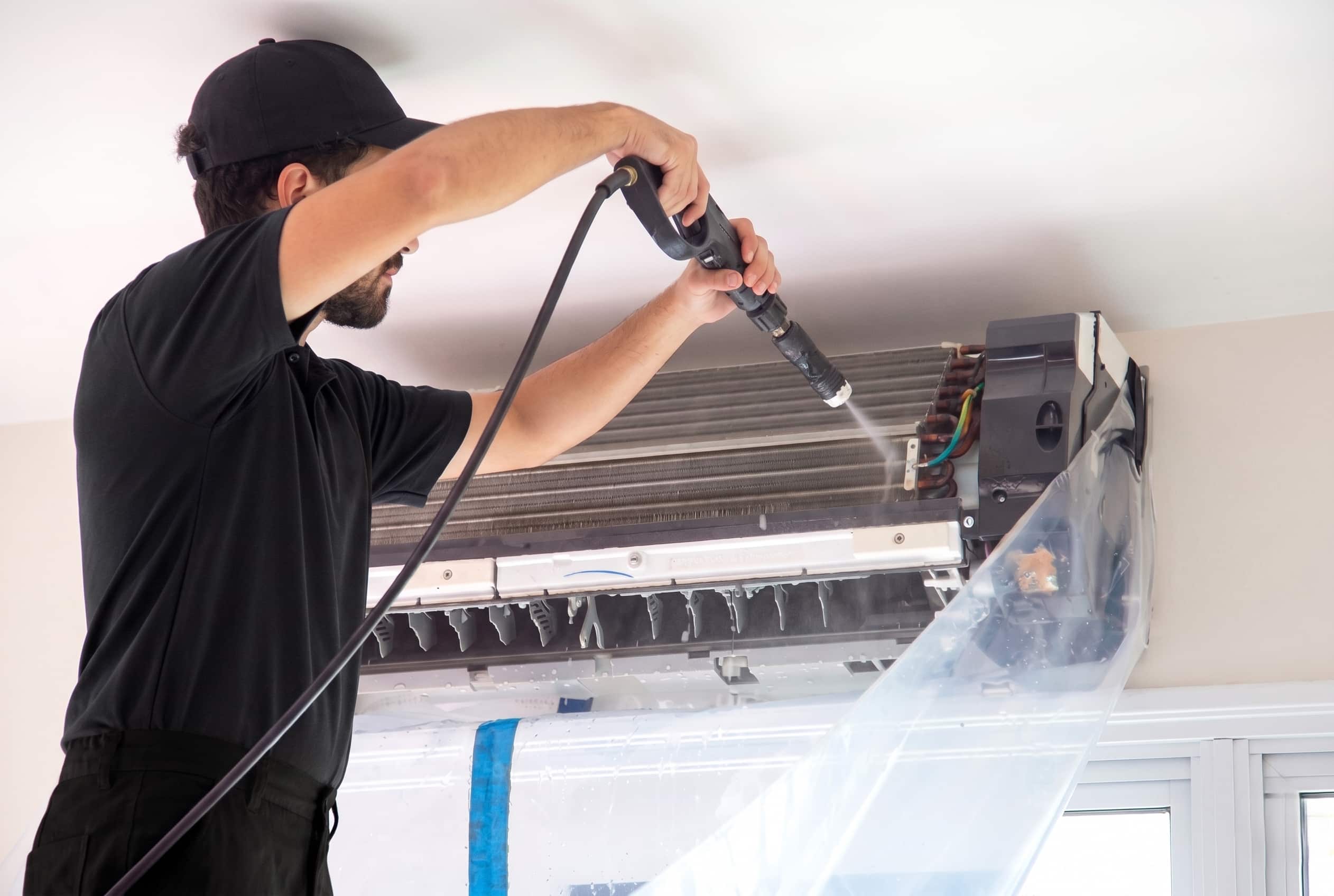 Heat pump technician using a low-pressure wand to clean the internal coil of a wall-mounted unit, with the front panel removed and a protective wash bag fitted to contain water runoff.