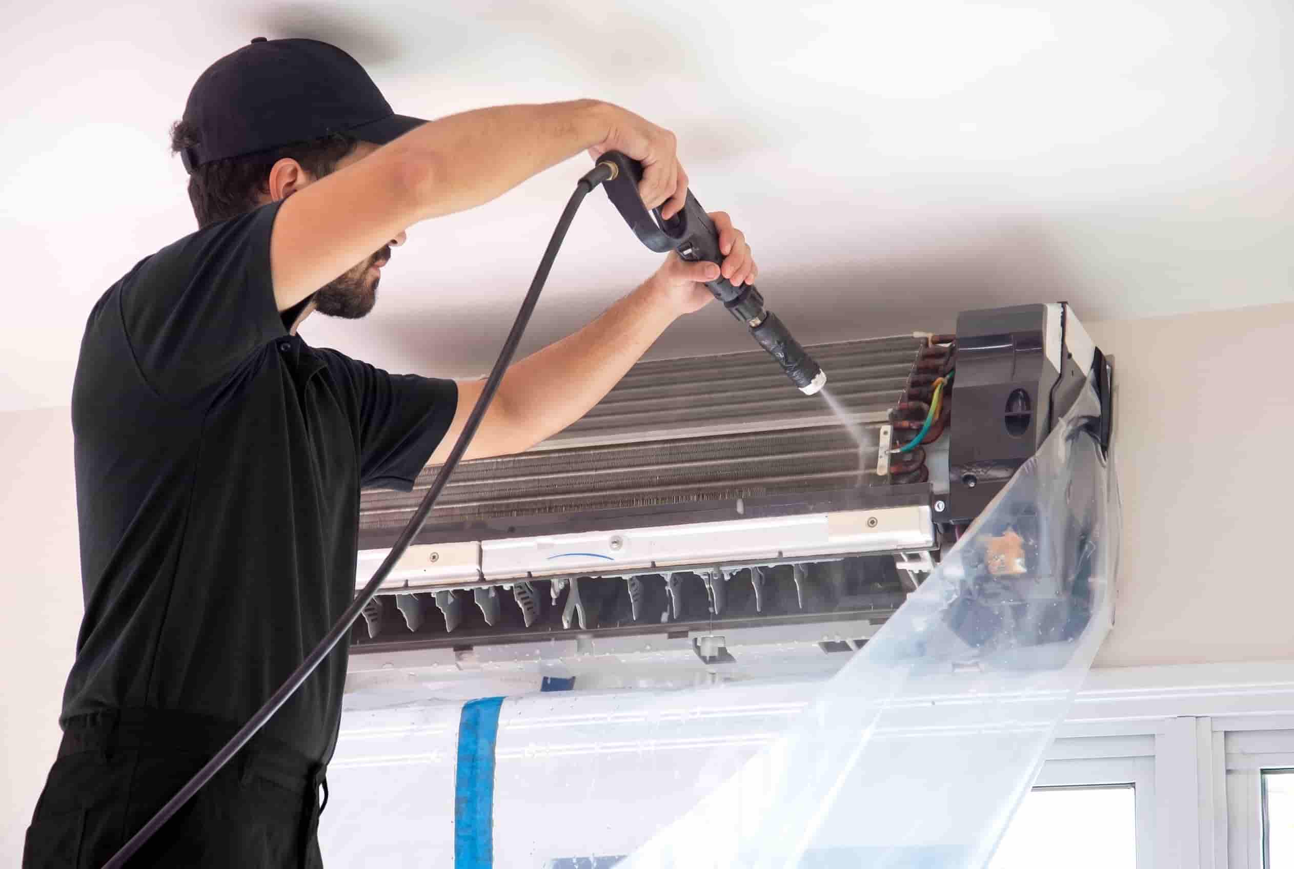 Heat pump technician using a low-pressure wand to clean the internal coil of a wall-mounted unit, with the front panel removed and a protective wash bag fitted to contain water runoff.