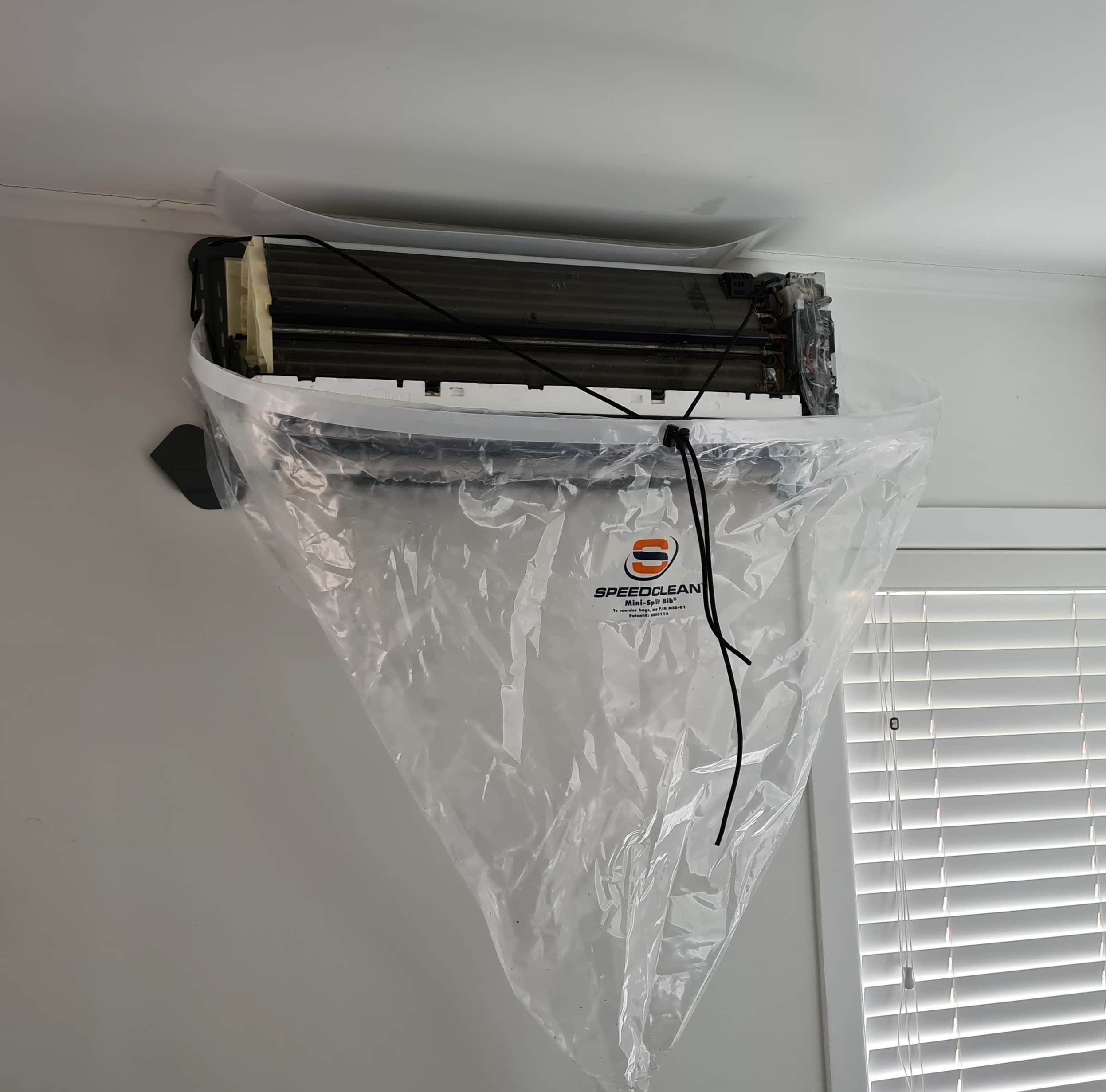 Wall-mounted heat pump indoor unit with front panel removed and a cleaning bib fitted to contain water runoff during a professional coil flush service in an Auckland home.