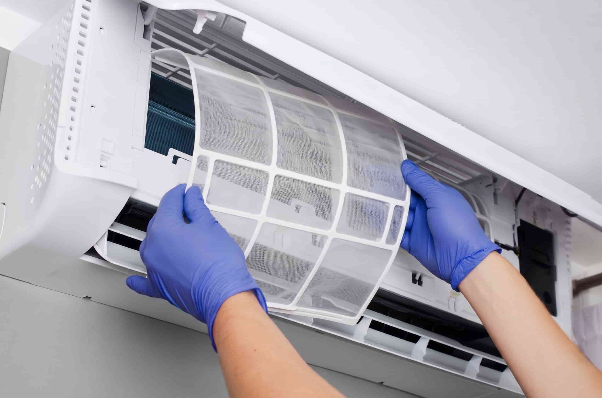 Person removing a heat pump filter from a wall-mounted indoor unit, showing the mesh filter frame and internal coil behind it.