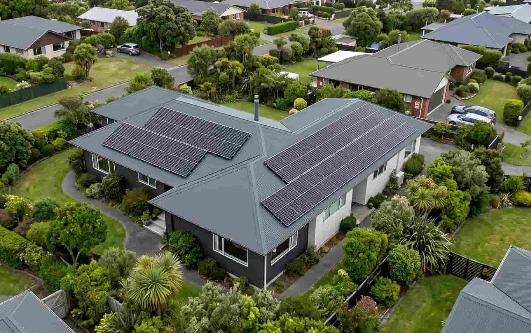 Aerial view of a New Zealand home with solar panels mounted on a dark grey metal roof, surrounded by green suburban gardens in Auckland.