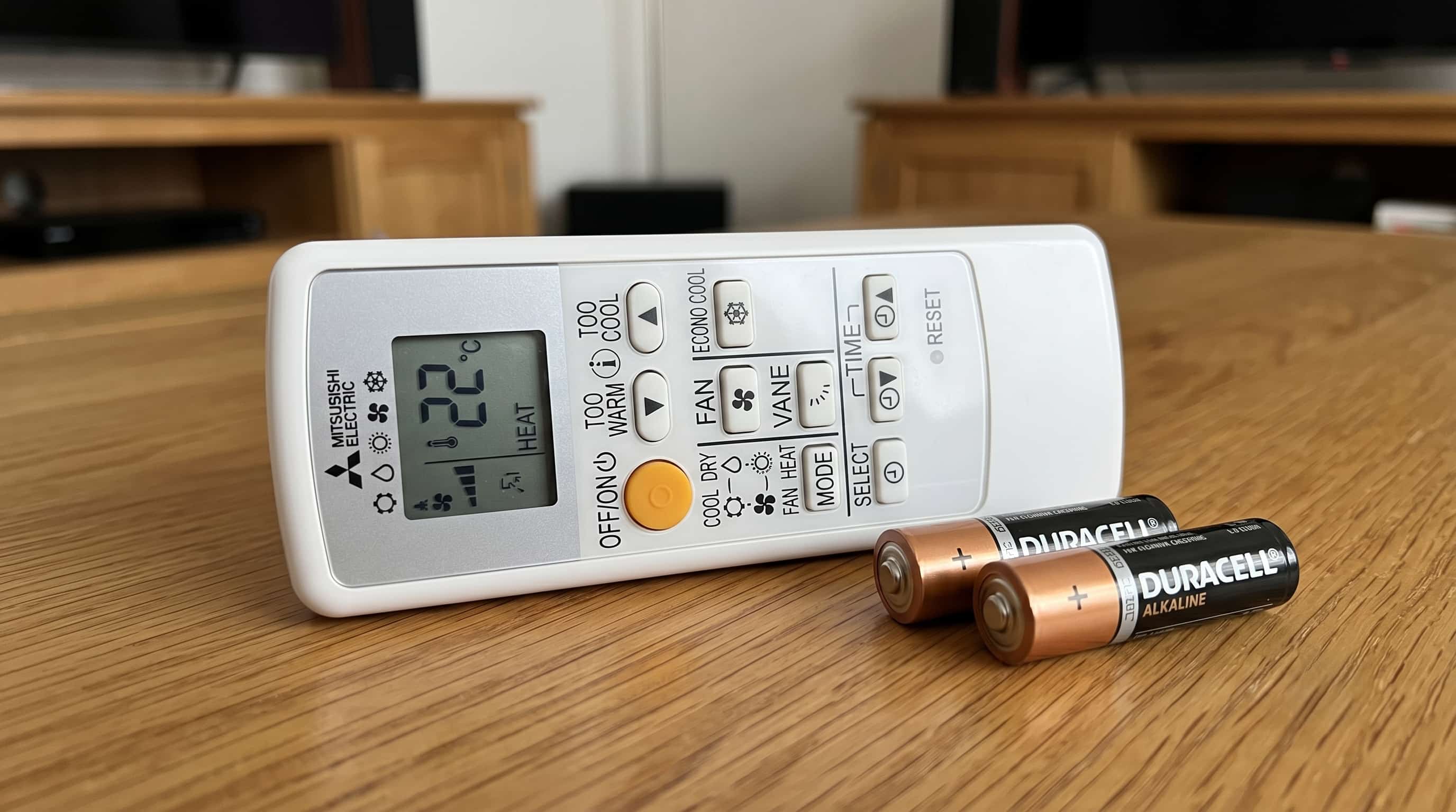 Mitsubishi Electric heat pump remote with two AA alkaline batteries on a wooden surface, showing the RESET button