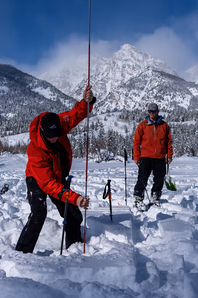 A mountain rescuer in full winter gear tending to an injured person lying in the snow on a steep alpine slope