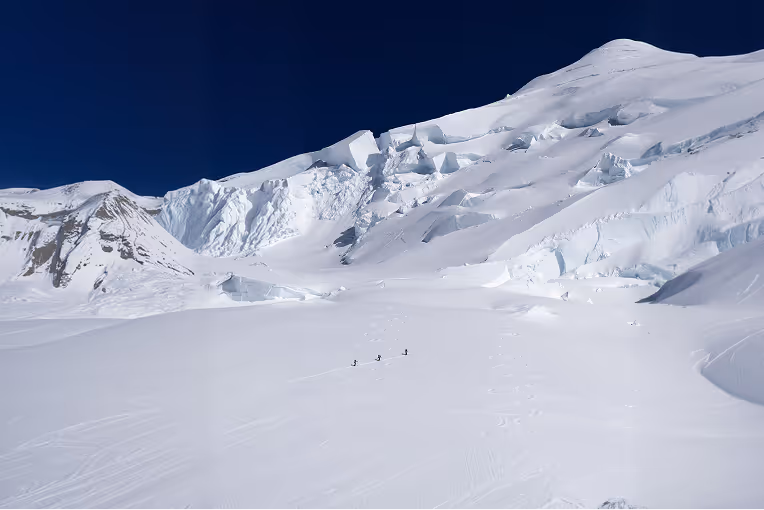 A group of mountaineers climbing a steep snowy ridge in single file, with dramatic mountain peaks and a cloudy sky in the background