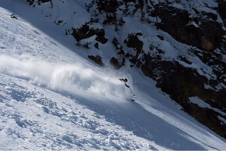 A skier in a red jacket descending a steep, untracked powder slope in a mountain backcountry setting