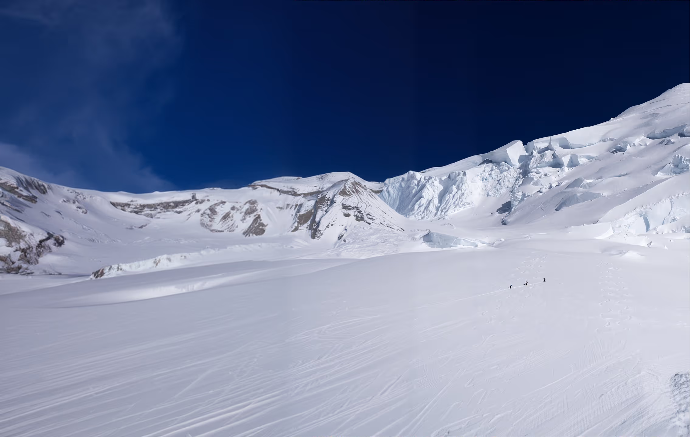 A group of mountaineers climbing a steep snowy ridge in single file, with dramatic mountain peaks and a cloudy sky in the background