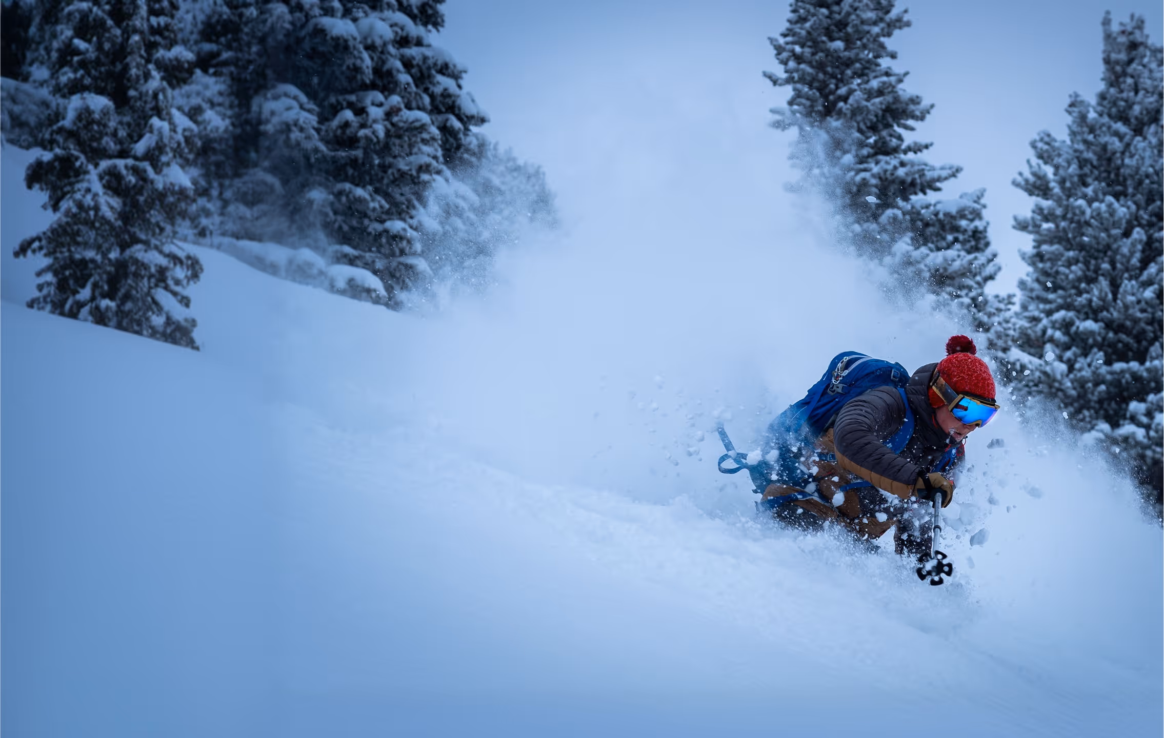 Skier with a red helmet skiing down a slope with powdery snow an pine trees in the background