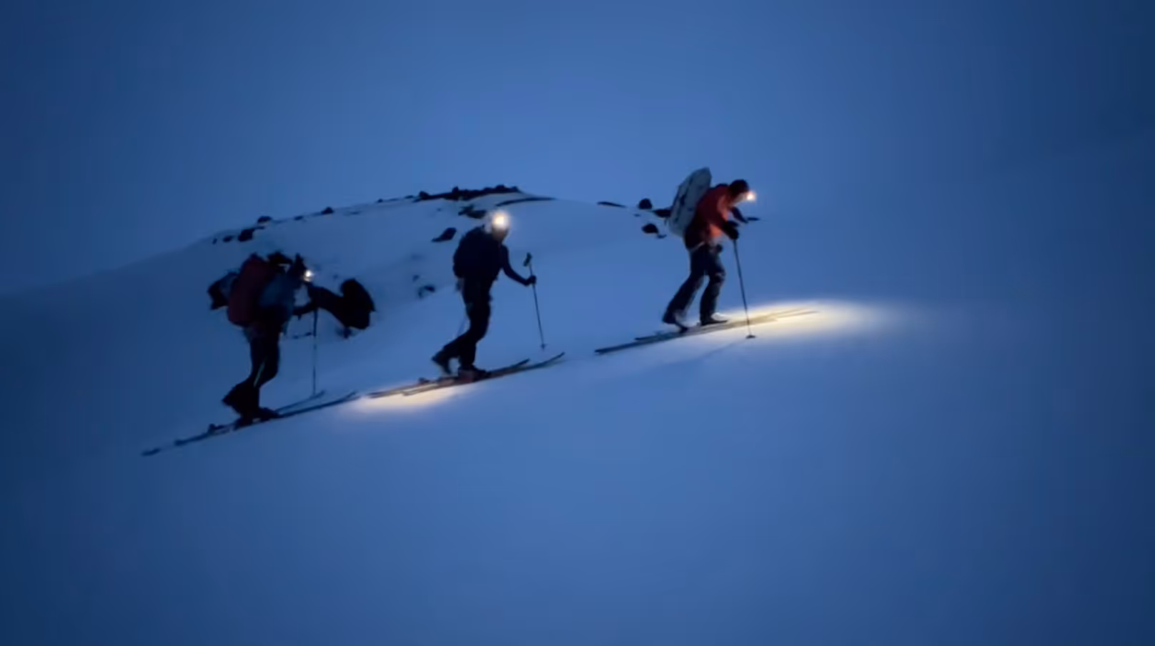 Three mountain climbers ascending a hill in snowy conditions in the dark with headlamps on