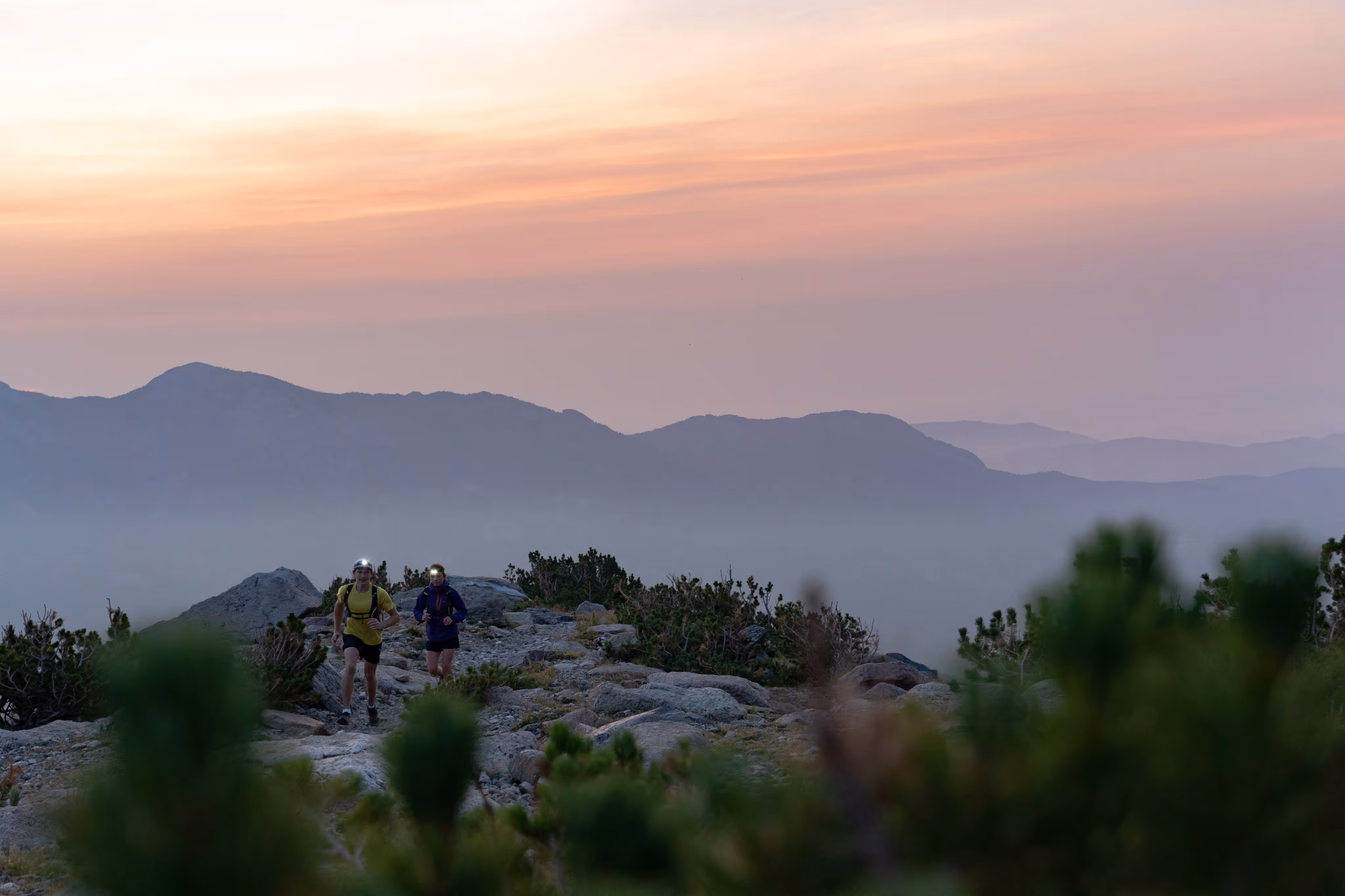 2 mountain athletes running on a mountainous trail with a scenic mountain panorama behind them