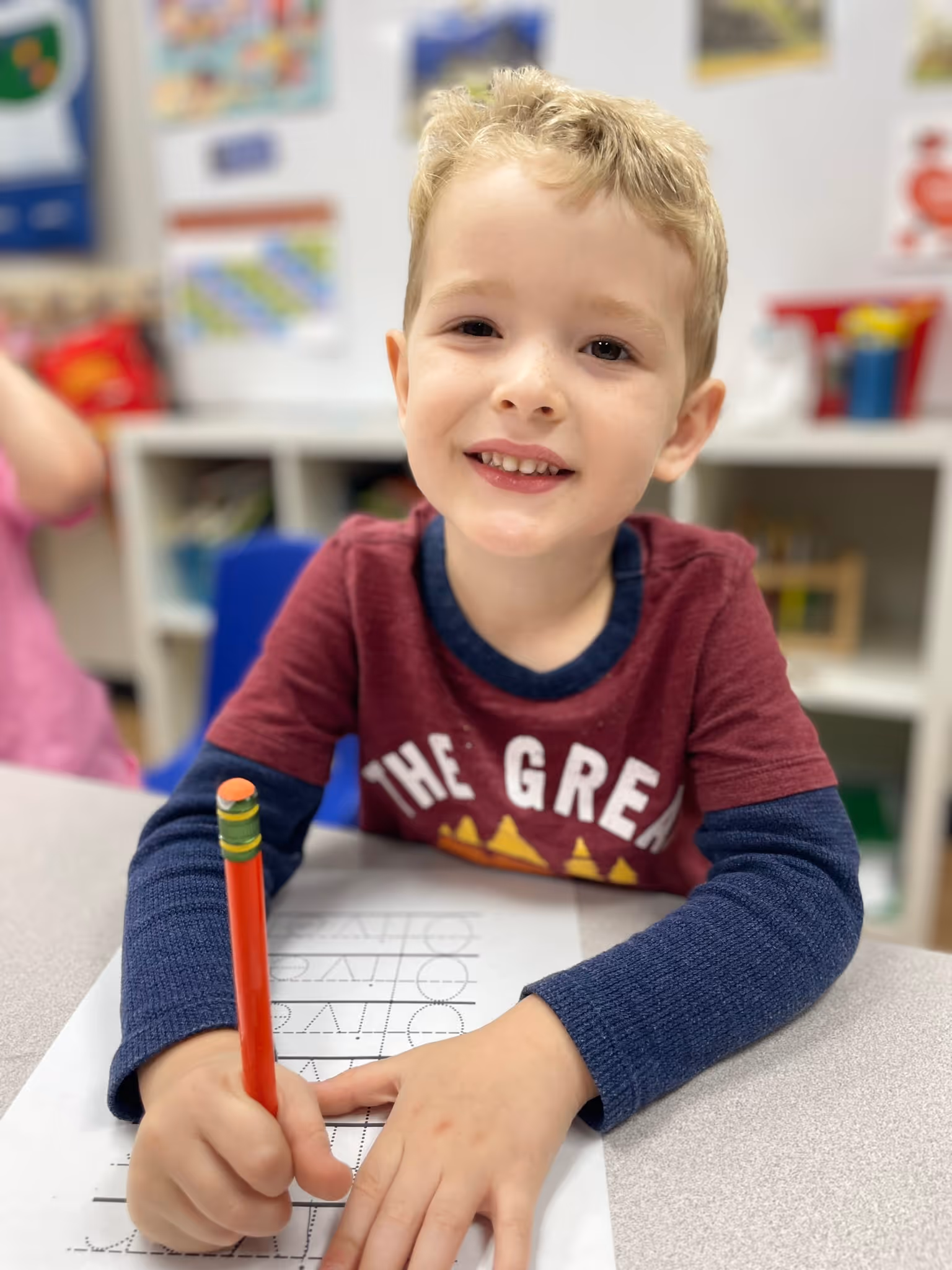 Young boy in a classroom smiling while holding a pencil and practicing writing on lined paper.