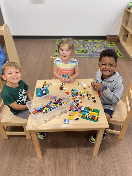 Three young children sitting at a wooden table playing with colorful building blocks and smiling at the camera.