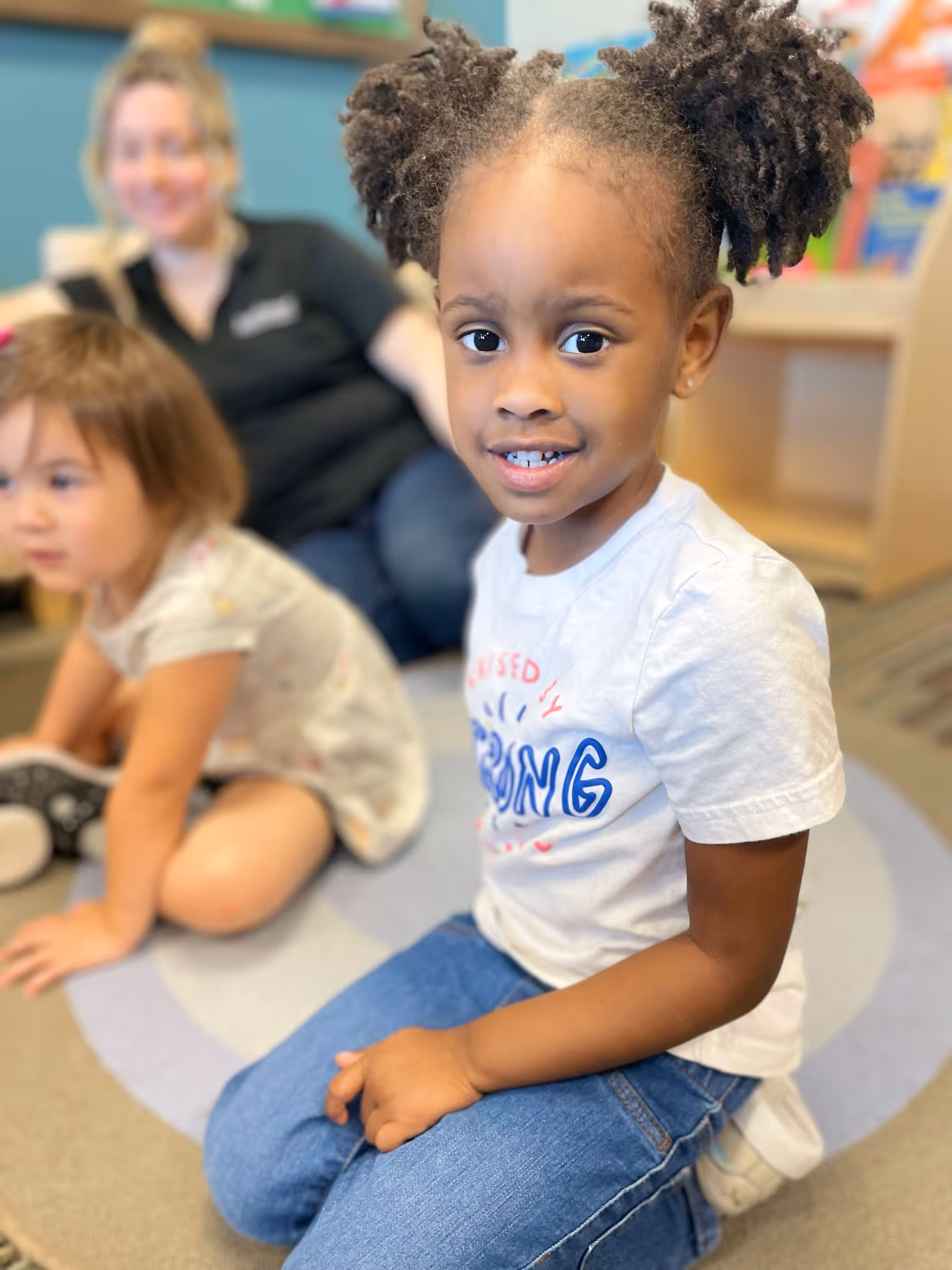 Young girl with pigtails wearing a white shirt and jeans sitting on a rug, with another child and an adult blurred in the background.
