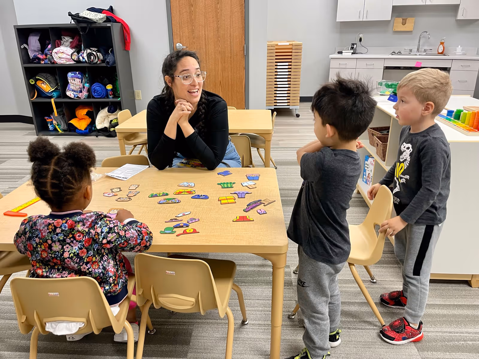 Teacher sitting at a table engaging with three children in a classroom with educational puzzle pieces on the table.
