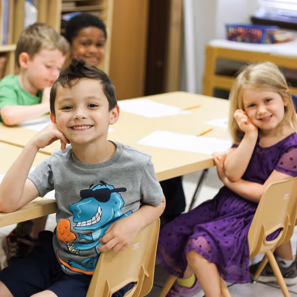 Four young children sitting at a classroom table smiling, with two children in the front and two blurred in the background.
