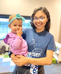 Smiling woman wearing glasses and a blue t-shirt holding a baby in a purple outfit with a green headband indoors.