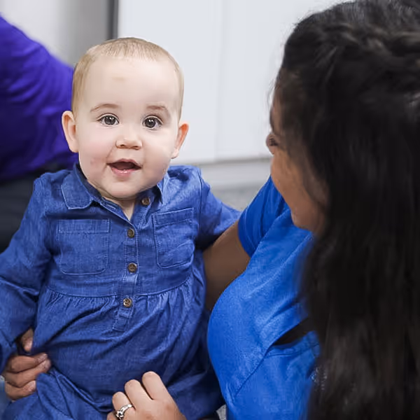 A woman in a blue shirt holding and smiling at a happy baby wearing a denim dress.