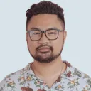 Headshot of a man with short black hair, beard, glasses, and a floral patterned shirt against a light gray background.