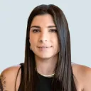 Portrait of a woman with long straight dark hair, light skin, and tattoos on her left shoulder, wearing a black top and a gold necklace, against a light blue background.