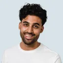 Smiling man with short curly hair and a beard wearing a white shirt against a light background.